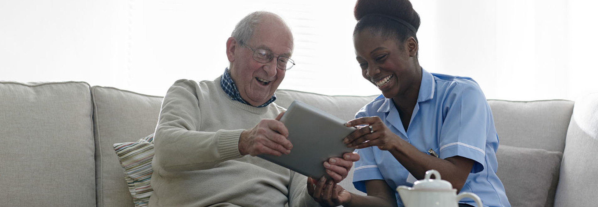 Happy senior man and home nurse sitting on sofa using digital tablet - Indoors