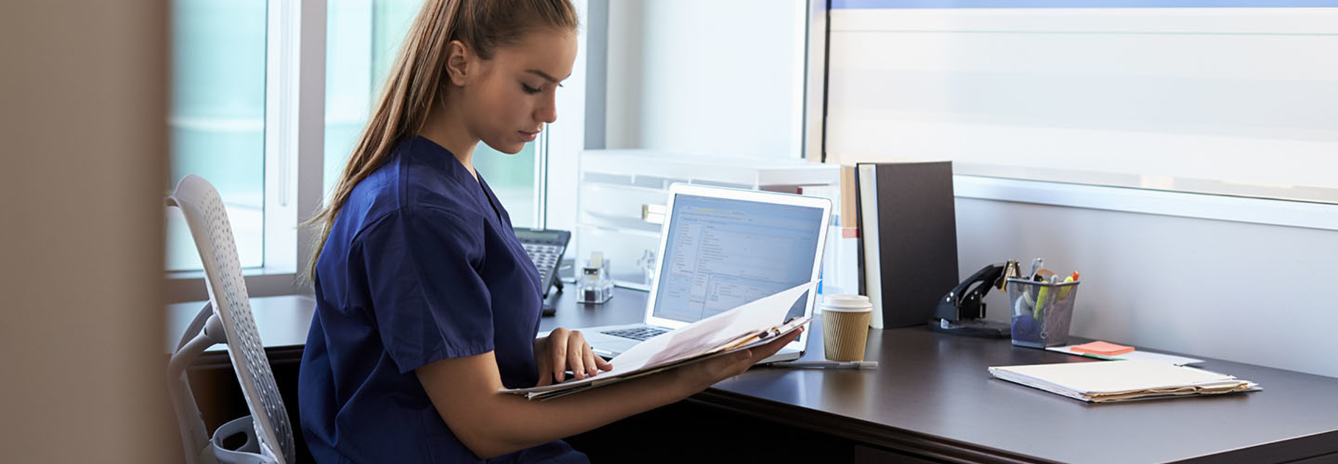 Nurse Wearing Scrubs Working At Desk In Office