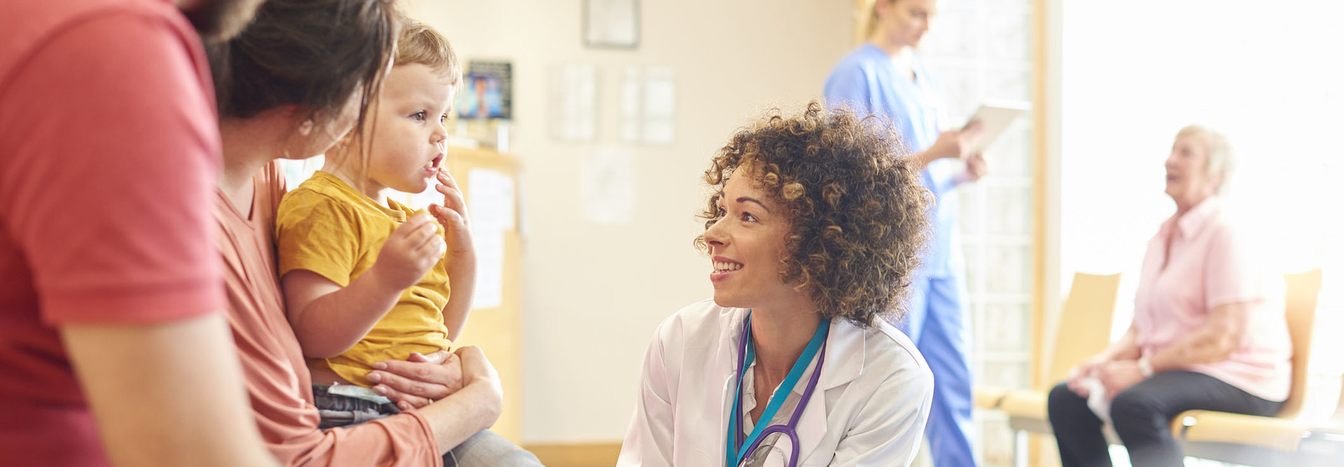 A young family sits talking to the doctor. The toddler is sat on his mother’s knee as the doctor kneels down to talk to him