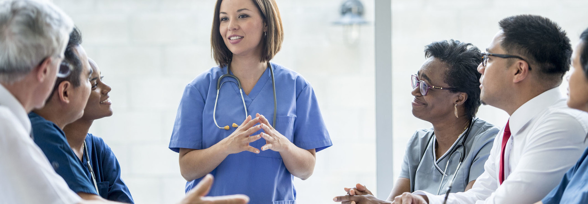 A multi-ethnic group of medical staff are indoors in a hospital. They are wearing medical clothing. A Caucasian female doctor is giving a presentation to the others.
