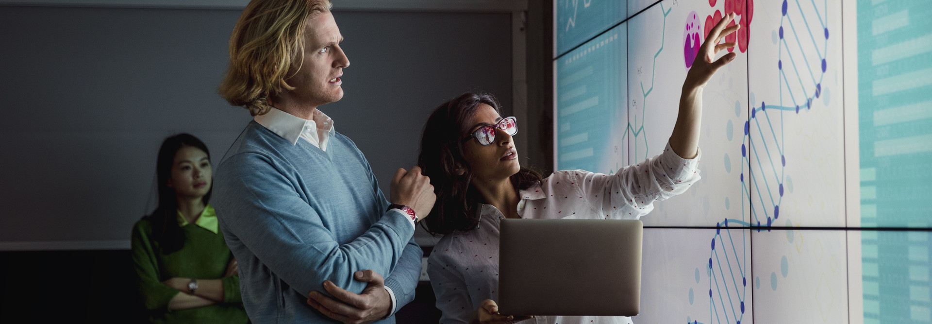 Group of business professionals in a dark room standing in front of a large data display screen with information.