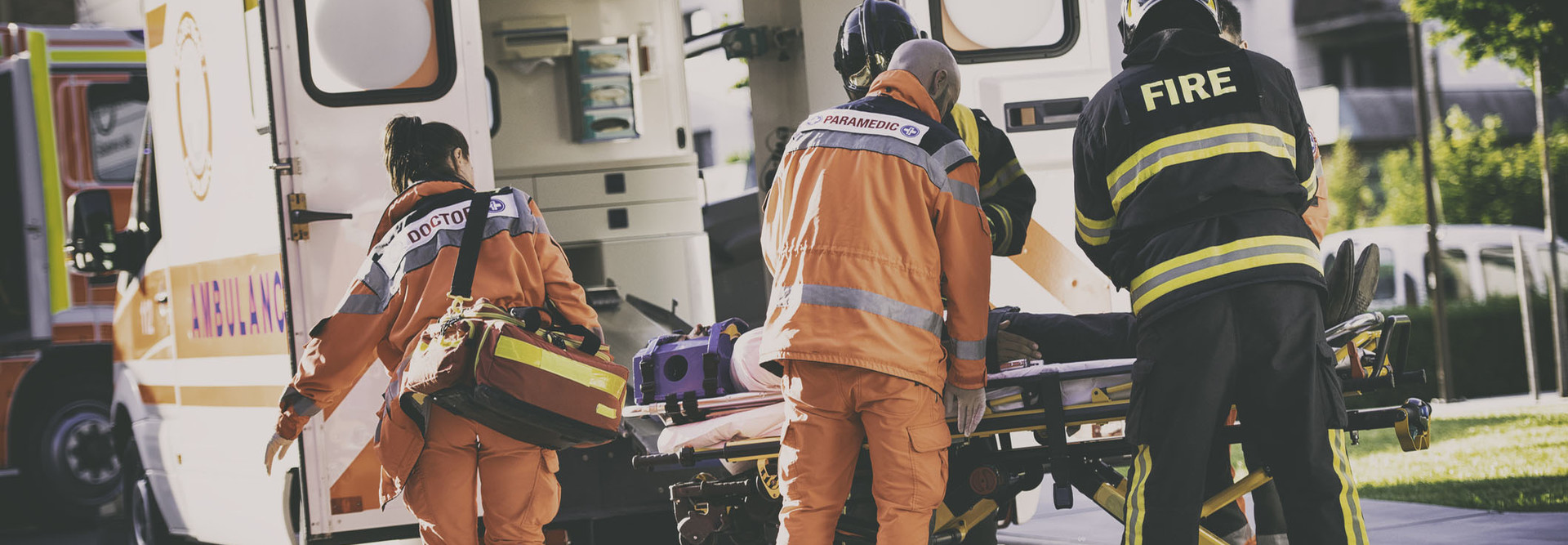 Two firefighters helping one female and one male paramedic to put a stretcher with injured person into ambulance vehicle.