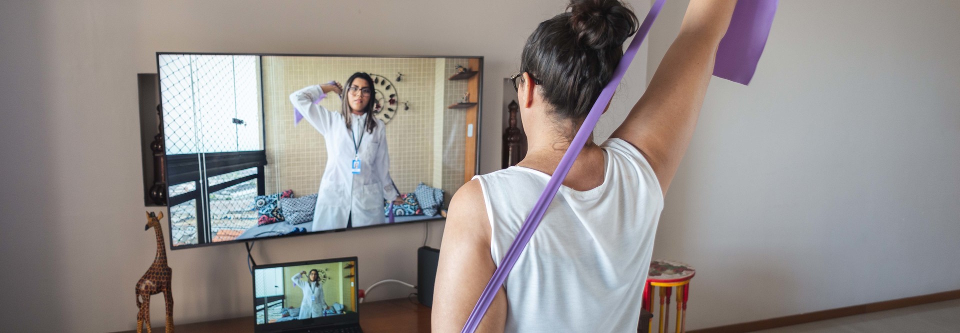 oman watching and copying exercises with a resistance band in her living room, guided by a physical therapist online