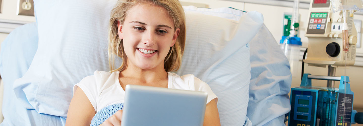 A Teenage Female Patient Relaxing In Hospital Bed With a Digital Tablet. She is Lying Down.