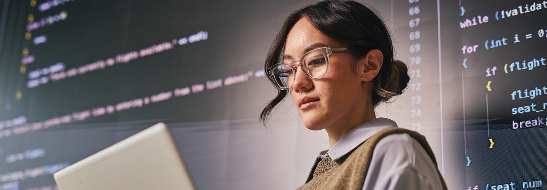 woman wearing glass holds laptop in front of wall of code