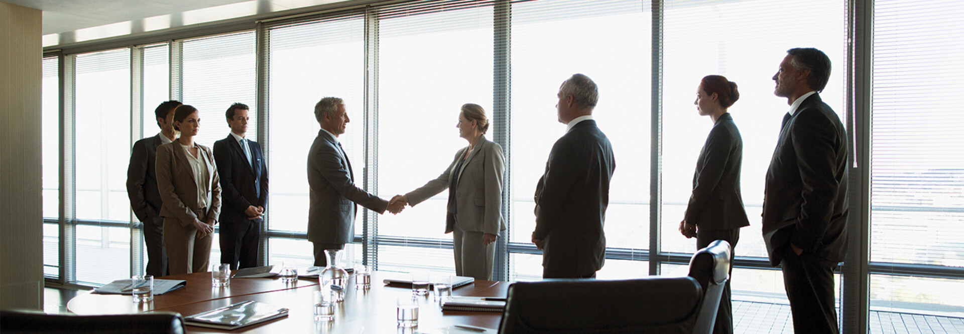 business people gather around conference table as leaders shake hands