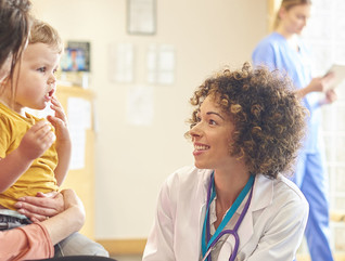A young family sits talking to the doctor. The toddler is sat on his mother’s knee as the doctor kneels down to talk to him