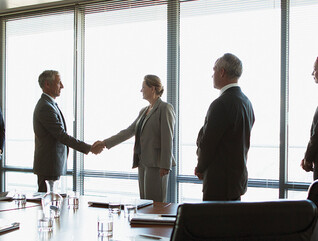 business people gather around conference table as leaders shake hands