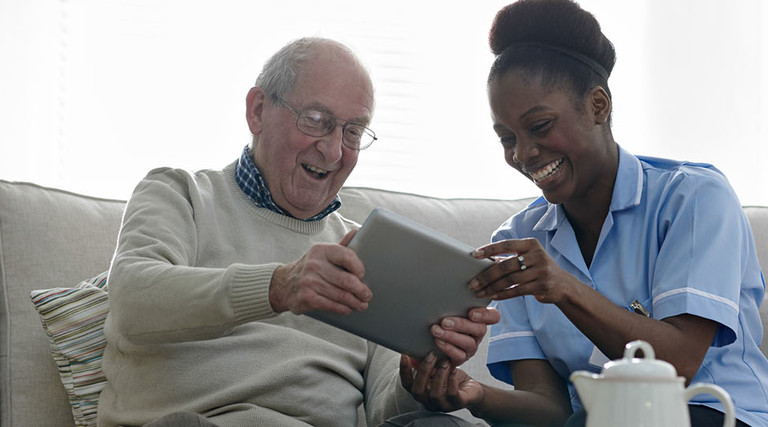 Happy senior man and home nurse sitting on sofa using digital tablet - Indoors