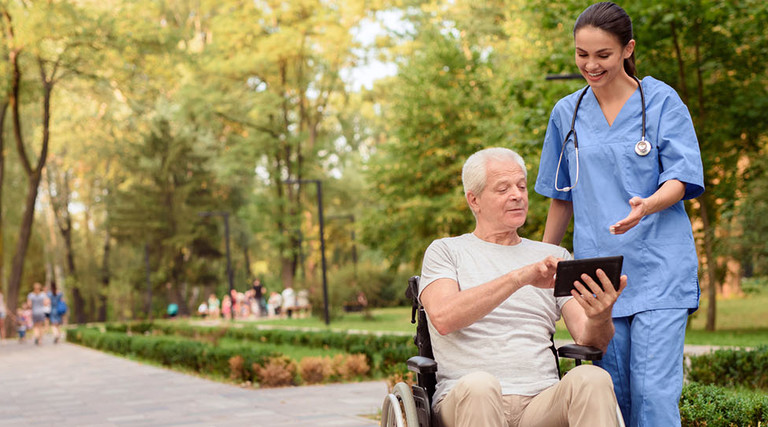 An old man sitting in a wheelchair shows the nurse something on his tablet