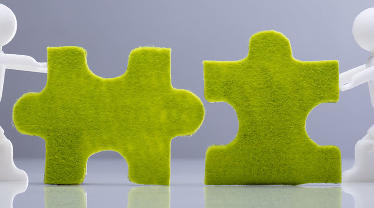 Side View Of Two Miniature Human Figures Solving Green Jigsaw Puzzles On Grey Background