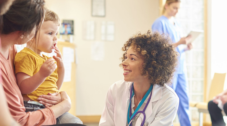 A young family sits talking to the doctor. The toddler is sat on his mother’s knee as the doctor kneels down to talk to him