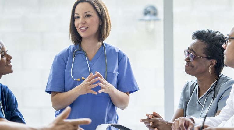 A multi-ethnic group of medical staff are indoors in a hospital. They are wearing medical clothing. A Caucasian female doctor is giving a presentation to the others.