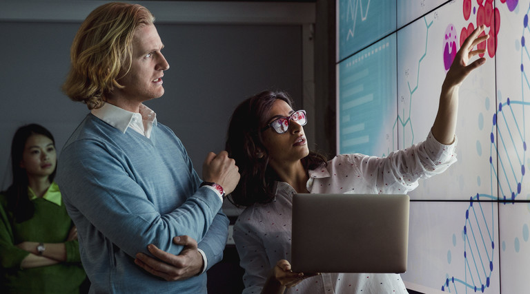 Group of business professionals in a dark room standing in front of a large data display screen with information.