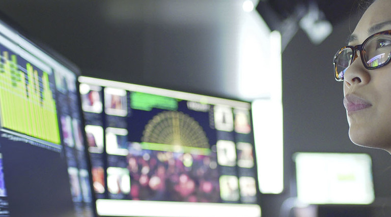Close up stock image of a young asian woman sitting down at her desk where she’s surrounded by 3 large computer monitors displaying out of focus images 