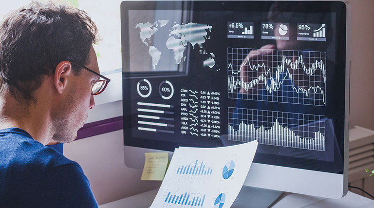 Man at desk looking at computer screen and files with charts and graphs.