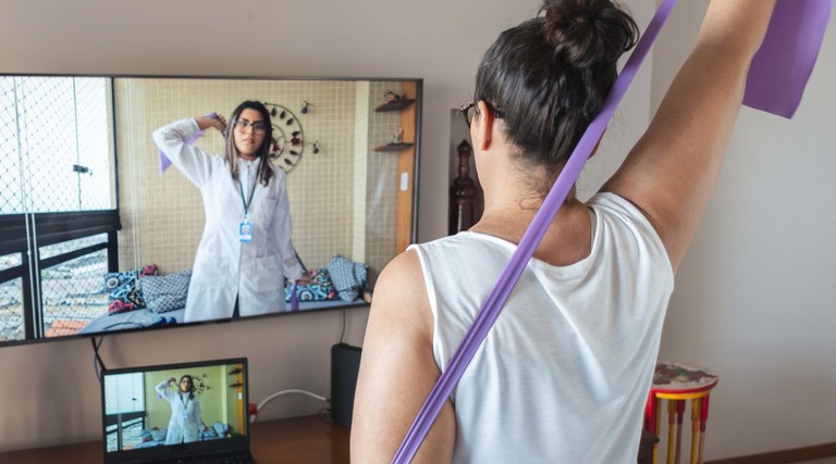 oman watching and copying exercises with a resistance band in her living room, guided by a physical therapist online