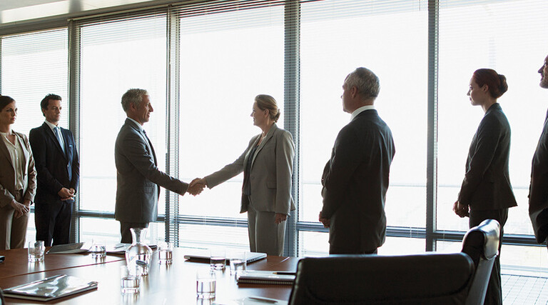 business people gather around conference table as leaders shake hands