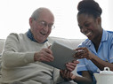 Happy senior man and home nurse sitting on sofa using digital tablet - Indoors