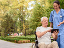 An old man sitting in a wheelchair shows the nurse something on his tablet