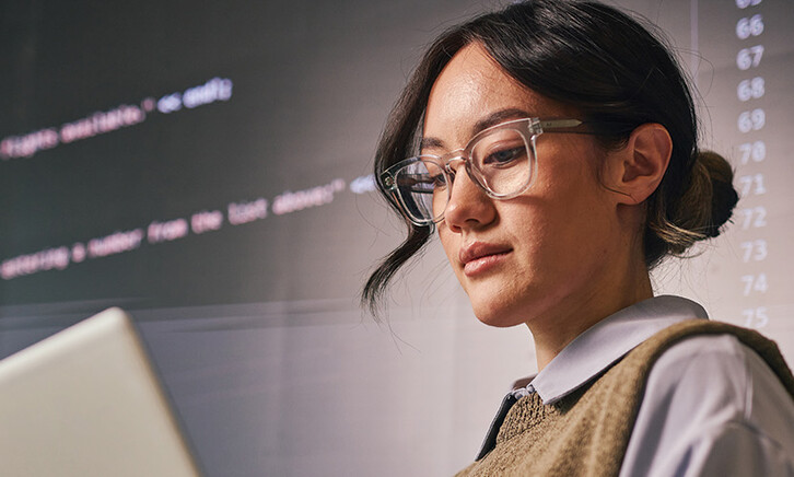 woman wearing glass holds laptop in front of wall of code