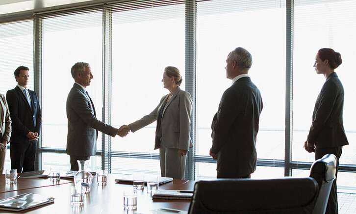 business people gather around conference table as leaders shake hands