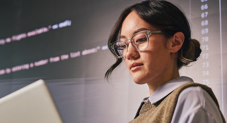 woman wearing glass holds laptop in front of wall of code