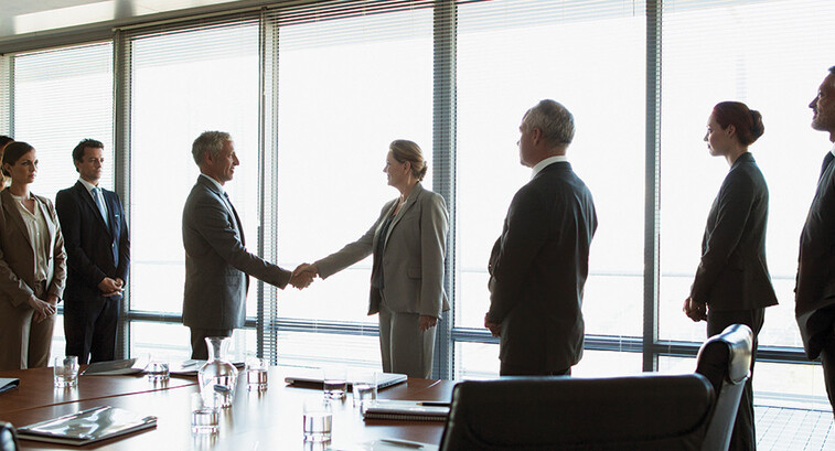 business people gather around conference table as leaders shake hands
